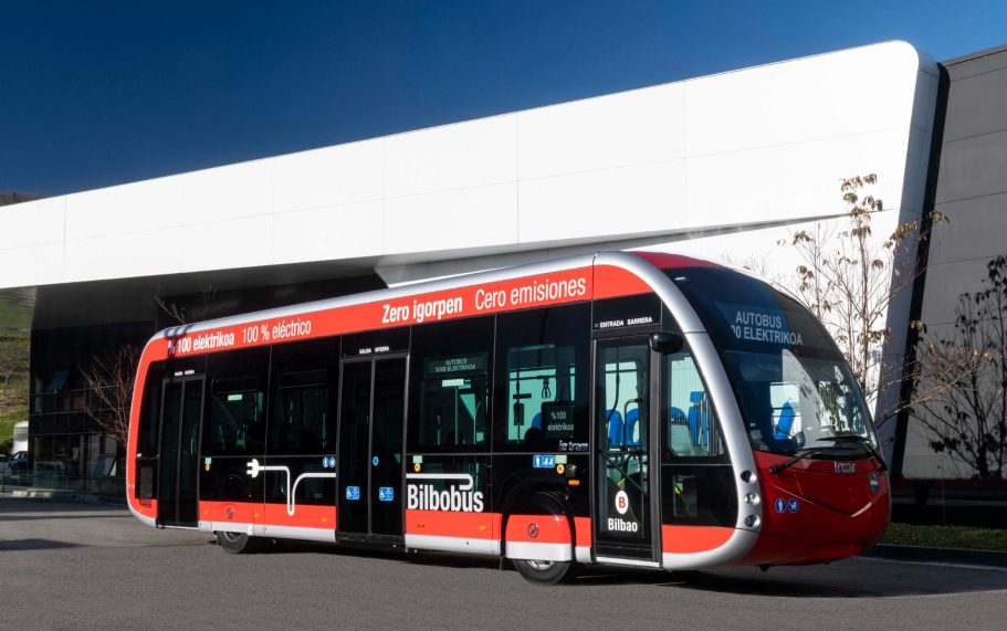 Autobús moderno de transporte público de color rojo y blanco, estacionado frente a un edificio.
