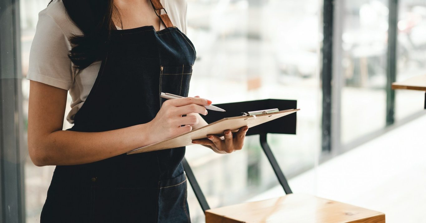 Persona con delantal negro tomando notas en una libreta en un entorno luminoso.