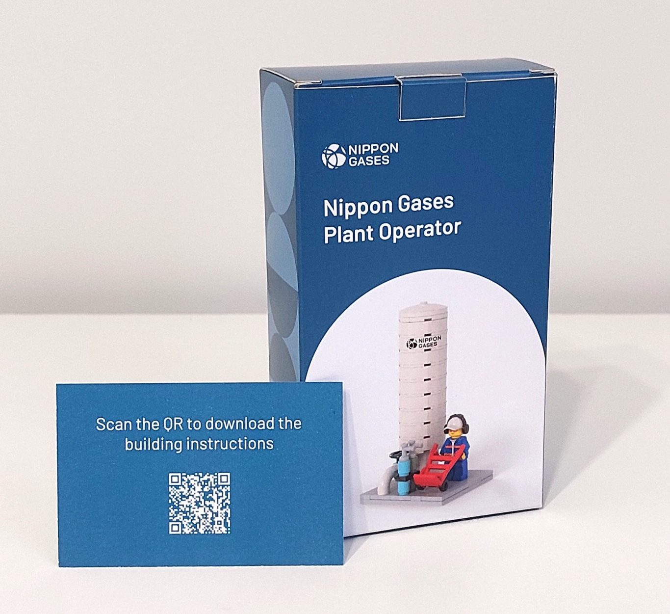 Caja de juego "Nippon Gases" con una torre y figuras de operarios, junto a una tarjeta QR.