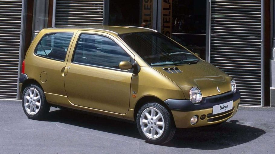 Coche pequeño de color dorado, modelo clásico, estacionado frente a un edificio moderno.